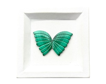 May include: Two carved malachite pieces shaped like butterfly wings, featuring deep green banded patterns and a ribbed texture, displayed together on a square white tray with a textured border and a smooth, recessed center area.