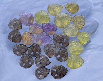 May include: A collection of small, carved gemstone fish beads in various colors including translucent yellow, light purple, and smoky gray. Each piece features detailed scale patterns and fin textures, arranged in a loose cluster on a white surface.