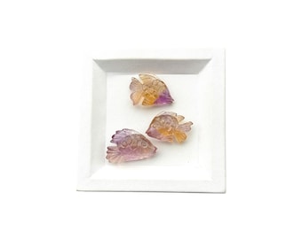 May include: Three small, translucent fish-shaped carvings made of multi-colored stone, featuring gradients of purple, yellow, and clear tones. Each piece displays detailed fin and scale textures, arranged on a square white plate with a textured border.