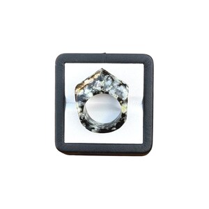 May include: A unique ring with a geometric design. The ring is made of a speckled black, white, and gray stone. The ring is displayed in a black square box with a white interior.