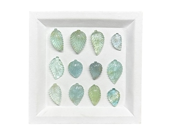 May include: A collection of twelve translucent, pale green and blue leaf-shaped gemstone carvings arranged in a grid on a white square display tray. Each individual stone features detailed, etched vein patterns and a small loop at the top.
