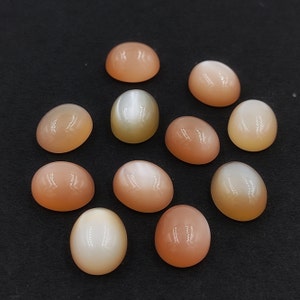 May include: A collection of ten oval-shaped moonstone cabochons in shades of white, peach, and pink. The stones have a smooth, polished surface and a slightly iridescent sheen.