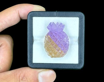 May include: A carved gemstone pineapple figurine featuring a dual-tone color gradient of translucent purple amethyst and golden yellow citrine, displayed inside a square black jewelry box with a white velvet insert.