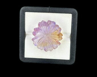 May include: A carved gemstone ring featuring a flower-shaped design with translucent petals transitioning from light purple to a warm golden yellow hue, presented in a square black display box with a white interior lining and a central slot.