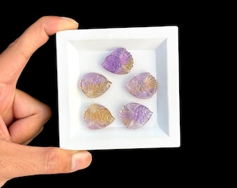 May include: Five small, carved gemstone fish figurines arranged in a white square tray. Each piece features a translucent gradient of purple and yellow hues, with detailed etched scales and fins visible on the surface of the polished stone carvings.