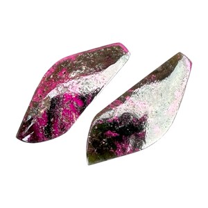 Pode incluir: Duas pedras preciosas polidas e alongadas com um esquema de cores rosa e prata vibrante. As pedras têm uma aparência em camadas, com tons de rosa intercalados na superfície prateada. As pedras preciosas estão sobre um fundo branco.