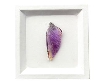 May include: A carved gemstone pendant shaped like a wing, featuring a gradient of translucent purple amethyst transitioning into a warm amber tone at the base, displayed centered within a square white frame with a recessed border.
