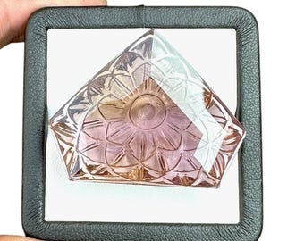 May include: A faceted gemstone with a floral design, displayed in a black case. The gem features a gradient of pink and peach hues, with intricate carvings that catch the light. The gem is held against a white background.
