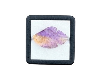May include: A carved gemstone fish figurine featuring a gradient of translucent purple and soft orange hues. The fish displays detailed etched lines representing scales and fins, presented inside a square black display box with a white interior.