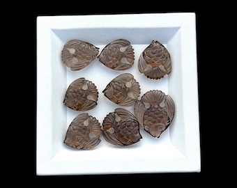 May include: Eight translucent, smoky brown, carved gemstone fish beads arranged in a white square tray. Each bead features detailed scale and fin engravings, with a small hole positioned near the top for threading, showcasing a polished finish.