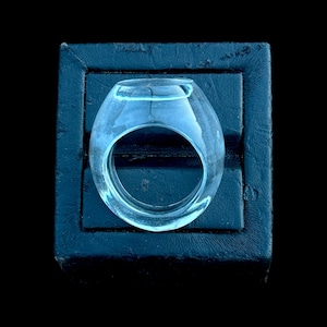May include: A transparent, pale blue glass ring with a rounded top and a large circular aperture. The ring is displayed on a dark blue, textured surface, providing a strong contrast. The ring's design is contemporary and minimalist.