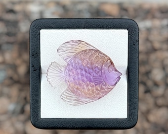 May include: A carved gemstone in the shape of a fish, featuring a gradient of translucent purple and orange hues. The carving displays detailed scale patterns on the body and defined lines on the fins and tail, presented in a square black display box.