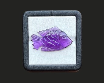 May include: A translucent purple amethyst gemstone carved into the shape of a fish. The carving features detailed etched lines representing scales, fins, and a tail. The fish is displayed inside a square black box with a white interior lining.