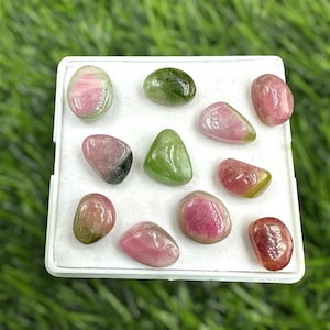 Puede incluir: Una colección de 10 piedras preciosas verdes, rosas y rojas pulidas. Las piedras son lisas y pulidas, y están dispuestas en una caja blanca.