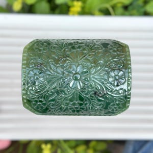 Può includere: Una gemma rettangolare verde giada con un intricato motivo floreale scolpito sulla sua superficie. Il design presenta fiori e foglie dettagliati, creando un aspetto ornamentale strutturato. I bordi della pietra sono leggermente arrotondati.