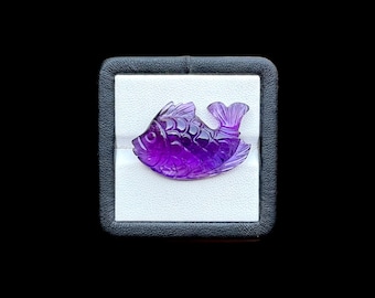 May include: A deep purple, translucent amethyst gemstone carved into the shape of a fish. The carving features detailed scale patterns, a defined eye, and a fanned tail. The gemstone is displayed inside a square, black-rimmed jewelry box.
