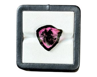 May include: A raw, triangular-shaped tourmaline gemstone featuring a vibrant pink center surrounded by a dark, black outer rim. The stone is set within a square, black-bordered display box with a white, padded interior insert for secure placement.
