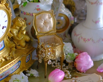 May include: A vintage gold clock with a white face and black hands sits on a table with a book, pink flowers, and a small gold jewelry box.