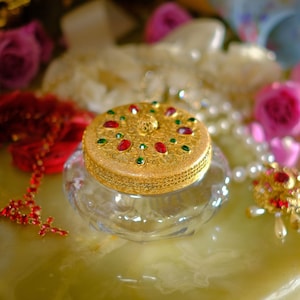 Puede incluir: Un tarro con tapa de oro y verde adornado con joyas con una base de vidrio transparente se encuentra sobre una superficie de mármol verde. El tarro está rodeado de perlas blancas, cuentas rojas y flores rosas.