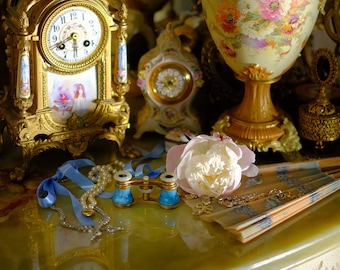 May include: A still life arrangement featuring a gold-toned antique clock, a vase filled with pink peonies, and a small decorative clock. Other items include a pearl necklace, blue ribbon, and a decorative fan. The scene is set on a reflective, ornate surface.