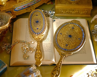 May include: Two antique gold-toned hand mirrors with blue jeweled accents and ornate designs, resting on an open book with a gold chain and other jewelry.