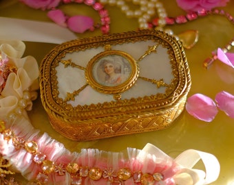 May include: A gold and white ornate jewelry box with a portrait of a woman inside. The box is sitting on a light green surface with a pink and white lace trim and a pearl necklace.