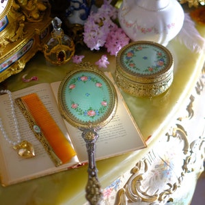 Pode incluir: Um conjunto de penteadeira vintage com um espelho de mão, uma pequena caixa de joias, um livro, um pente, um colar de pérolas e um vaso de porcelana com flores cor-de-rosa. Os itens estão dispostos em uma mesa com uma superfície decorativa.