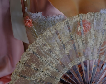 May include: A cream lace fan with a floral design is held in front of a cream corset. The fan has a dark brown frame. A pearl necklace and a pink rose are also visible. The overall theme is vintage and romantic.