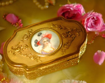 May include: A gold ornate jewelry box with a hinged lid and a small portrait of a woman in a white dress and a red hat. The box is surrounded by pink flowers, pearls, and a pink and white lace trim.