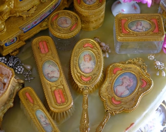May include: A set of antique gold-toned vanity brushes with pink accents and painted portraits. The brushes include a hairbrush, a clothes brush, and a hand mirror. The set is displayed on a marble surface.
