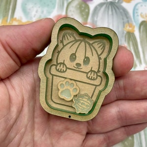 May include: A wooden cutout of a cartoon cactus wearing a pot with a paw print design. The cutout is light brown with a green outline and is held in a hand.