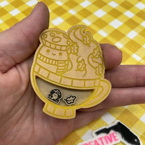 May include: A wooden cutout of a mug with a mushroom wearing a scarf and hat, surrounded by leaves. The mug is filled with a mushroom and leaves. The cutout is held in a hand. The text "THE NATIVE CREATIVE" is visible on a sticker in the background.