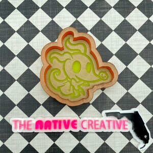 May include: A brown cookie cutter in the shape of a cartoon ghost with a pumpkin in its mouth. The cookie cutter is on a black and white checkered background. The text "THE NATIVE CREATIVE" is visible in pink on a white background.