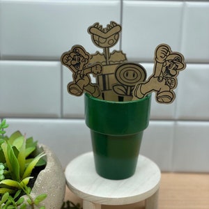 May include: A green ceramic planter with three cardboard cutouts of Mario characters: Mario, Luigi, and a Piranha Plant. The planter is on a small wooden stand. The text "THE native creative" is visible in the bottom right corner of the image.