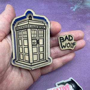 May include: A set of three wooden laser cut crafts. The first is a blue police box with the text "Police Box" on the side. The second is a small sign with the text "Bad Wolf" in black. The third is a white sticker with the text "The Native Creative" in pink and a black outline of the state of Florida.