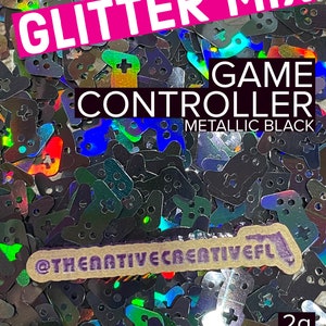 May include: A close-up of iridescent glitter shaped like game controllers. The glitter is labeled "Glitter Mix Game Controller Metallic Black" and "2g".