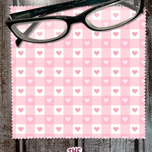 May include: Pink and white checkered microfiber cleaning cloth with a pink heart pattern. The cloth is folded in half and a pair of black eyeglasses is resting on top of it.