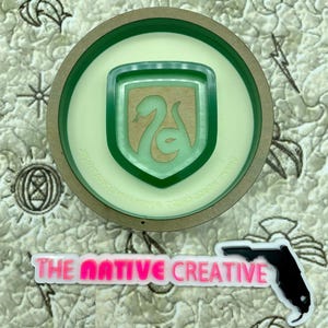 May include: A green and brown circular wooden coaster with a green snake emblem in the center. The emblem has the text "Slytherin" around the outside. The coaster is on a green and white patterned surface.