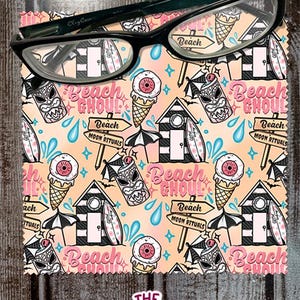 May include: Black-framed eyeglasses rest on a patterned cloth. The cloth features a beach-themed design with ice cream cones, tiki masks, beach huts, and the text "Beach Ghoul". The background is a light peach color.