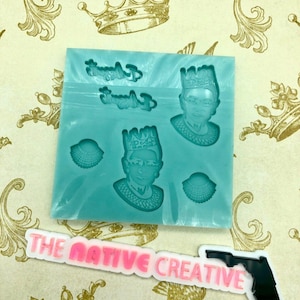 May include: A blue silicone mold with two impressions of Ruth Bader Ginsburg's face wearing a crown. The mold also has two smaller impressions of a crown. The text "The Native Creative" is visible in pink letters on a white background.