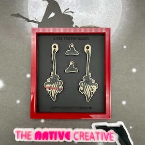 May include: A red framed display featuring witch-themed earrings and charms. The earrings are shaped like brooms with black and cream details. The charms are witch hats. The background includes a witch silhouette and the text "THE NATIVE CREATIVE".