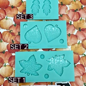 May include: Three sets of teal silicone molds for crafting, each featuring autumn-themed shapes like leaves and hearts. The molds have a leopard print pattern. The text "SET 1", "SET 2", and "SET 3" are visible. The background is a pumpkin pattern.