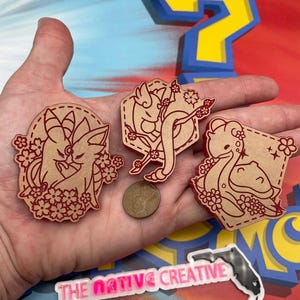 May include: Three wooden laser cut ornaments featuring different Pokemon characters. The ornaments are brown with red outlines and are decorated with cherry blossoms. The ornaments are held in a hand.