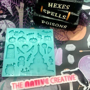May include: A teal blue silicone mold with various Halloween themed shapes, including a haunted house, pumpkins, bats, candy corn, a cauldron, a skull and crossbones, a tombstone, and a trick or treat bag. The mold is on a purple and orange patterned surface with a pink and white sticker that says "THE NATIVE CREATIVE".
