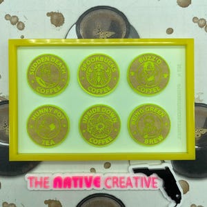 May include: A yellow-framed artwork featuring six circular coasters with pop culture-themed designs. The coasters are brown with neon green text and illustrations. The text includes "Sudden Death Coffee", "Spookbucks Coffee", and "Buzz'd Coffee". The artwork includes the text "The Native Creative".