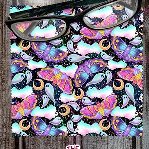 May include: Black-framed eyeglasses rest on a patterned fabric featuring colorful moths, ghosts, clouds, and crescent moons against a dark background. The fabric has the text "The Native Creative" in a pink and white design.