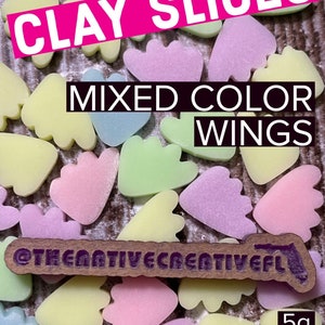May include: A close-up of a variety of pastel-colored clay slices shaped like wings. The clay slices are arranged on a wooden surface. The text "CLAY SLICES MIXED COLOR WINGS" is visible at the top of the image. The text "@THENATIVE CREATIVEFL" is visible at the bottom of the image. The text "5g THE NATIVE CREATIVE" is visible in the lower right corner of the image.