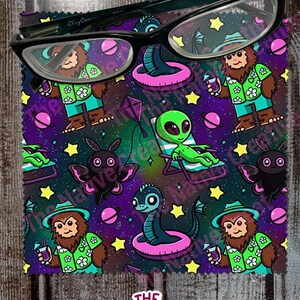 May include: A rectangular microfiber cleaning cloth featuring a repeating pattern of cartoon sasquatches in floral shirts, green aliens in lounge chairs, winged creatures, and planets on a dark purple background, topped with black eyeglasses.
