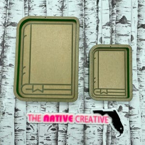May include: Two wooden book-shaped cutout shapes with a green border. The larger cutout is on the left and the smaller cutout is on the right. The cutouts are on a white background with a birch tree pattern.
