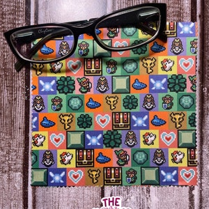 May include: A microfiber cleaning cloth with a colorful pixelated pattern featuring characters and items from the Legend of Zelda video game series. The cloth is folded in half and a pair of black eyeglasses are resting on top of it.